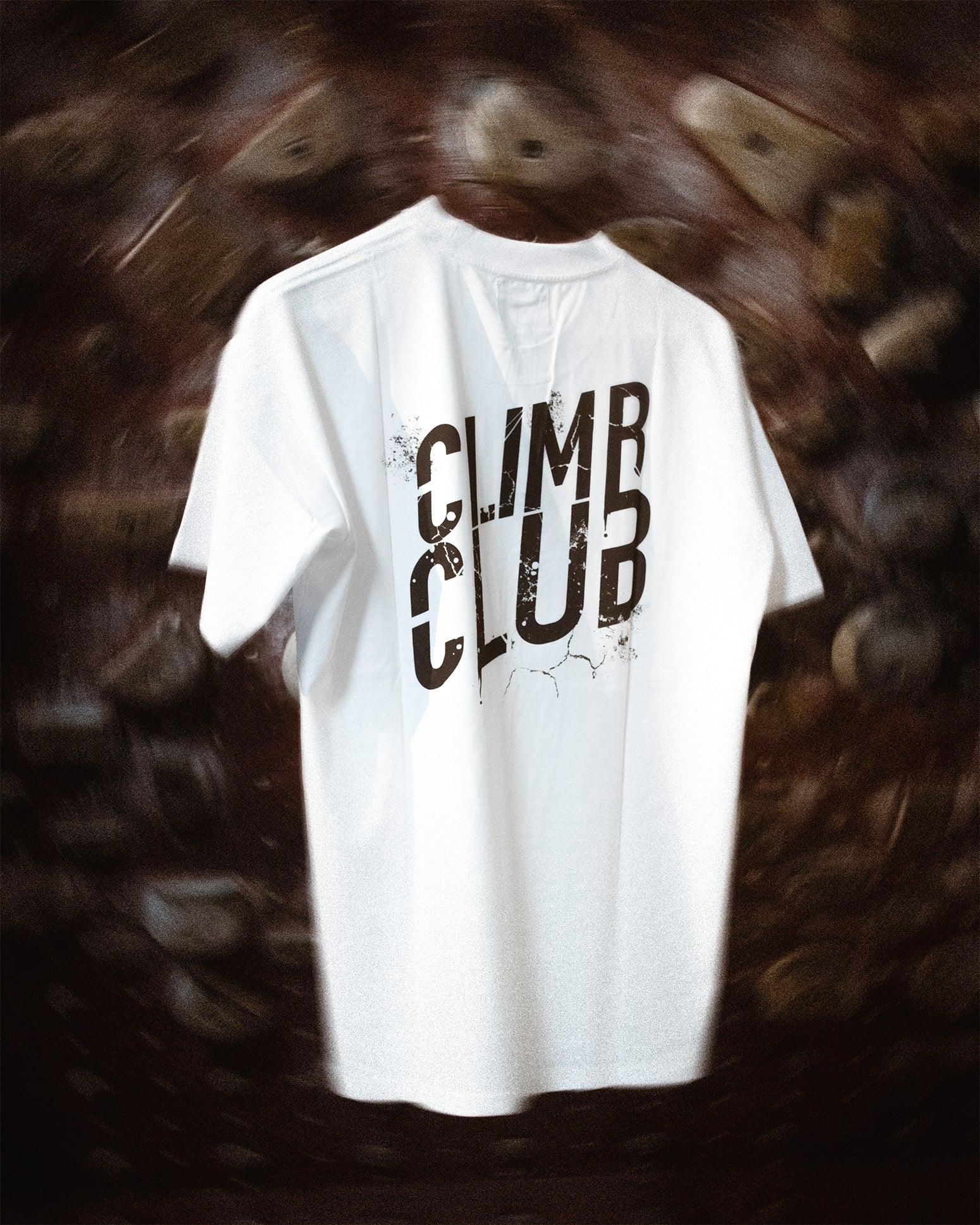 White Climb Club t-shirt with bold logo, hanging in front of an indoor climbing wall.